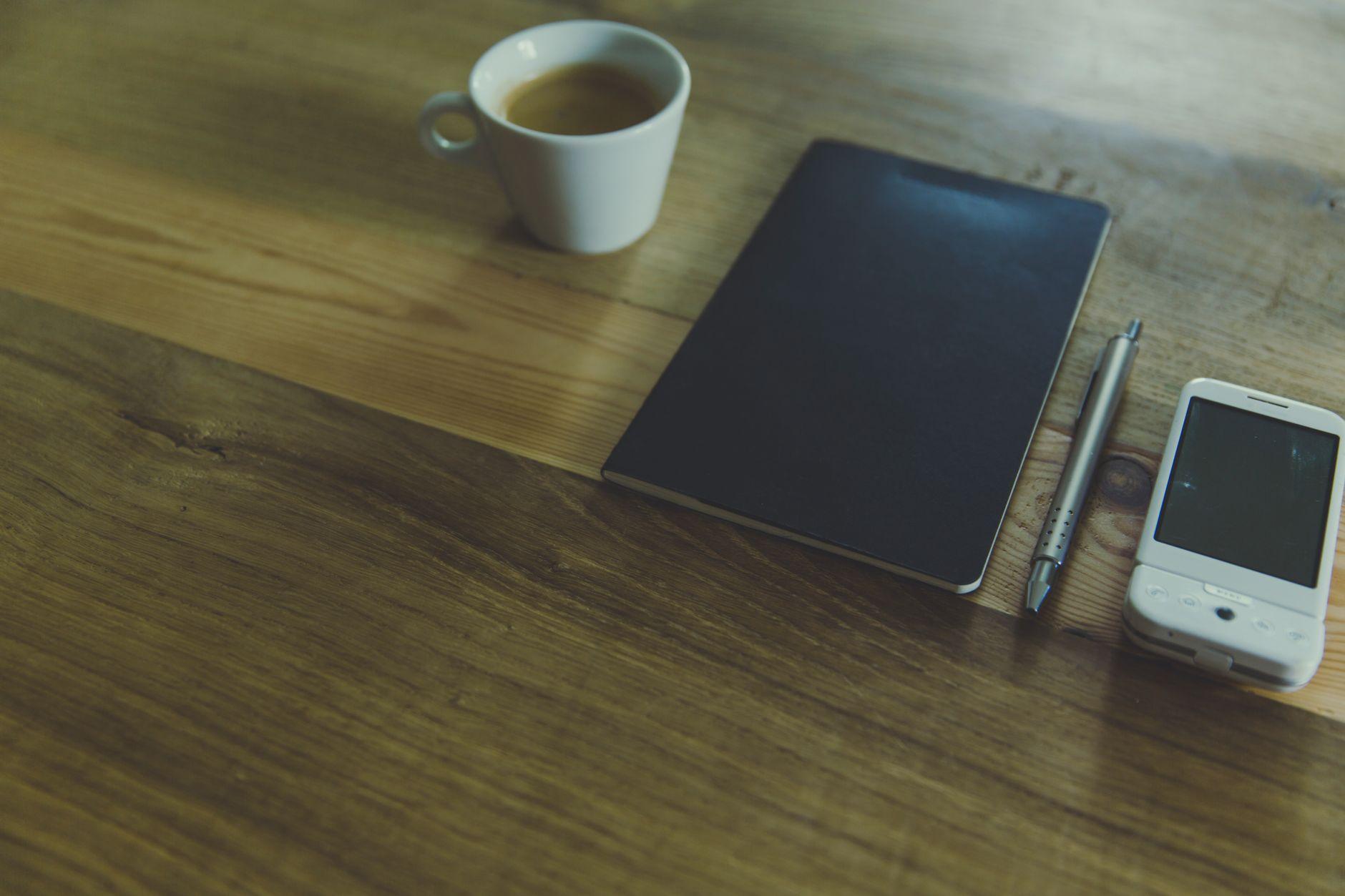 A notebook with handwritten notes and a coffee cup on a wooden desk in daylight